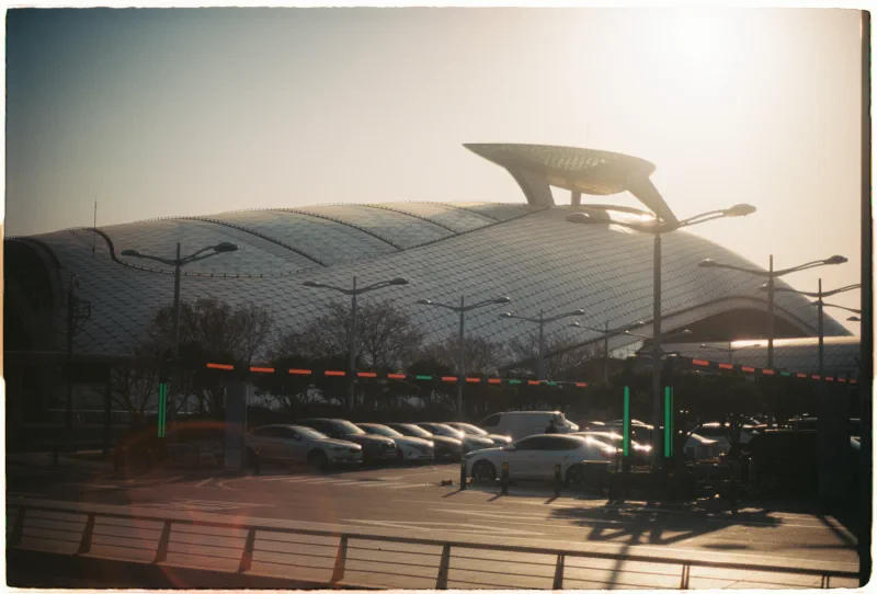 A curved airport terminal roof and parked cars sit in low golden sunlight