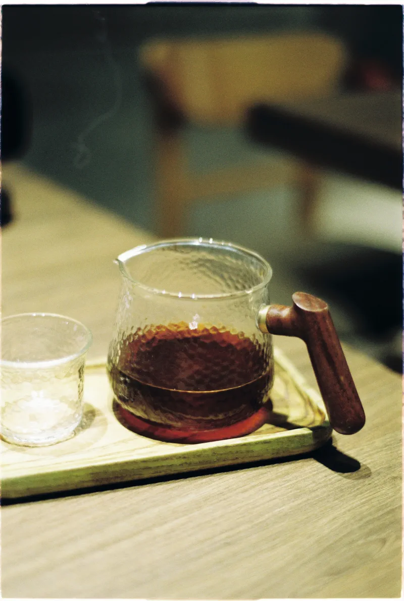 Amber tea rests in a hammered glass pot with a wooden handle on a pale tray