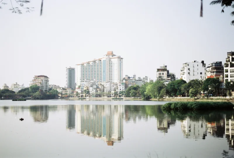 A pale Hà Nội skyline and trees reflect almost symmetrically in still lake water