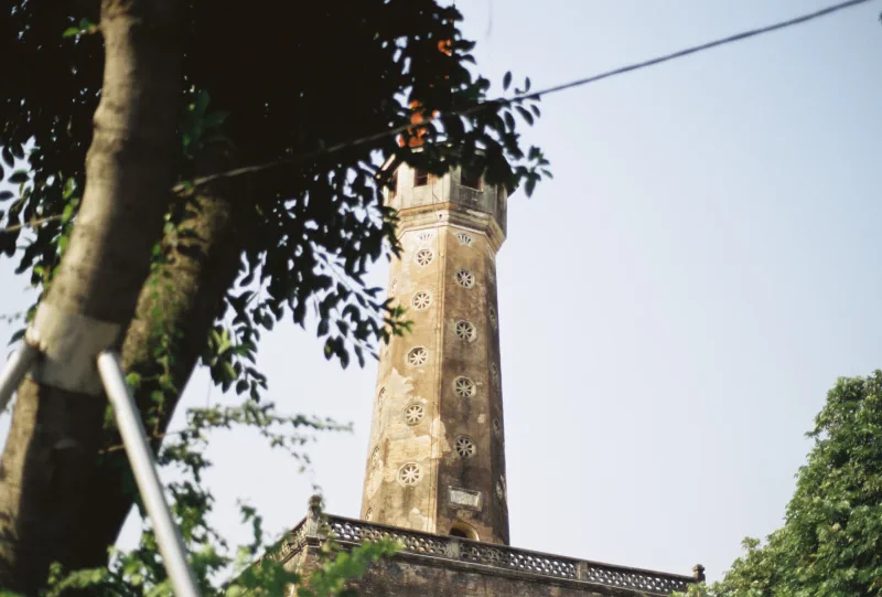 The weathered Hàng Đậu water tower rises behind a foreground tree in clear daylight