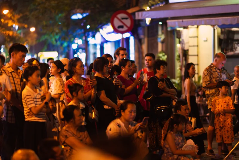 Children and adults gather shoulder to shoulder under warm streetlights to watch a tuồng performance in Hà Nội