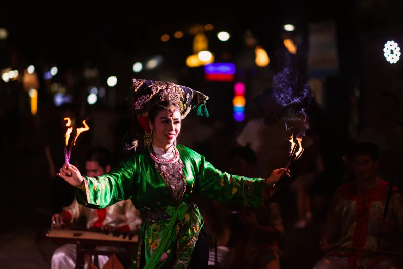 A performer in an embroidered green costume holds two flaming torches while musicians play behind her at night