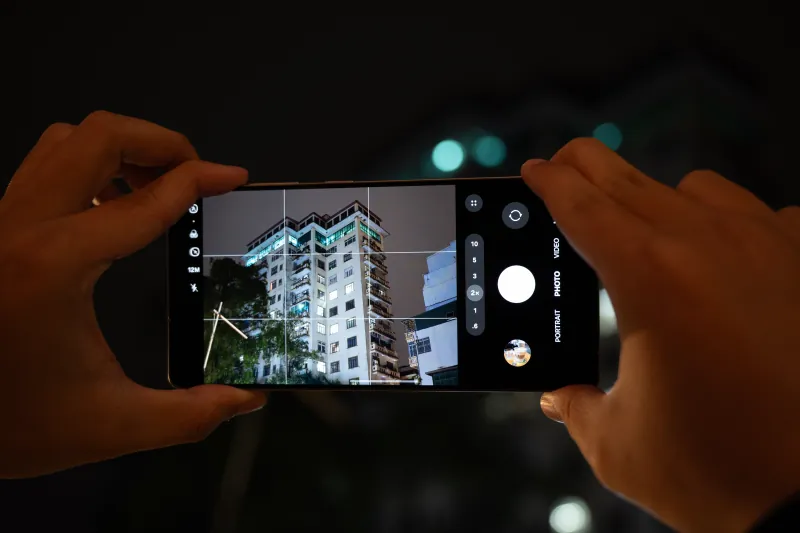 Two hands use the Galaxy S25 Ultra camera to frame a lit apartment building at night