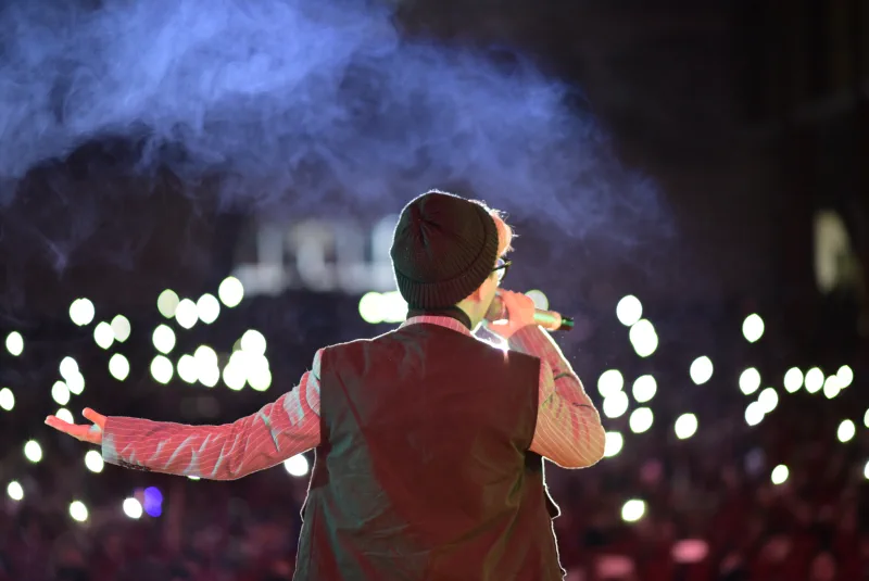 A vocalist in a beanie opens one arm toward an audience lit by phone screens
