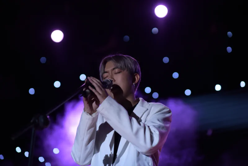A singer in a white suit closes his eyes at a microphone beneath purple lights